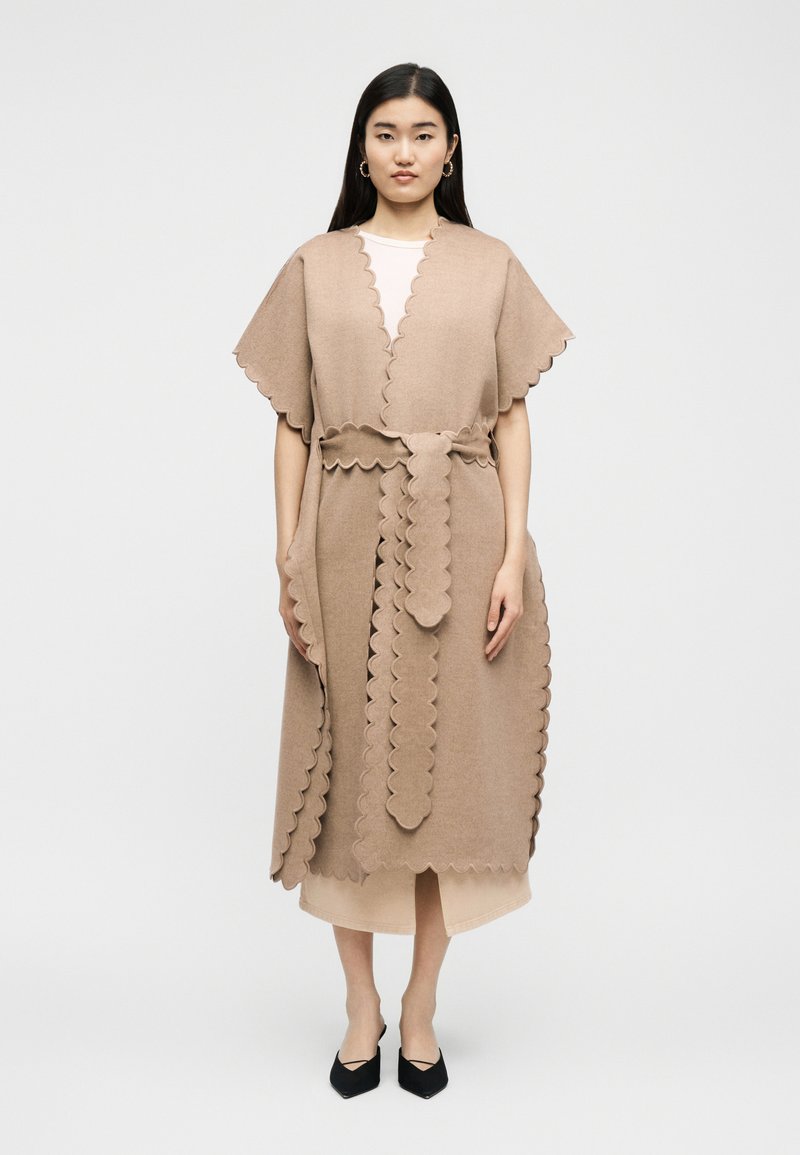 Frau trägt einen beige Wickelmantel mit geschwungenen Kanten und Gürtel, steht vor einem schlichten hellen Hintergrund, mit schwarzen spitzen Schuhen.