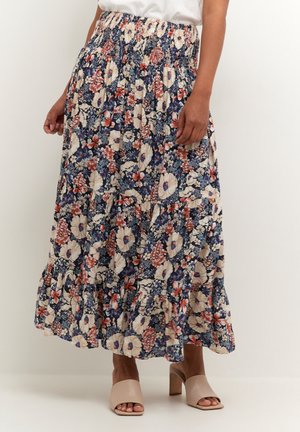 Femme portant une jupe midi à motif floral avec une taille smockée et des sandales à talons blocs ouvertes beige clair, debout contre un mur uni.