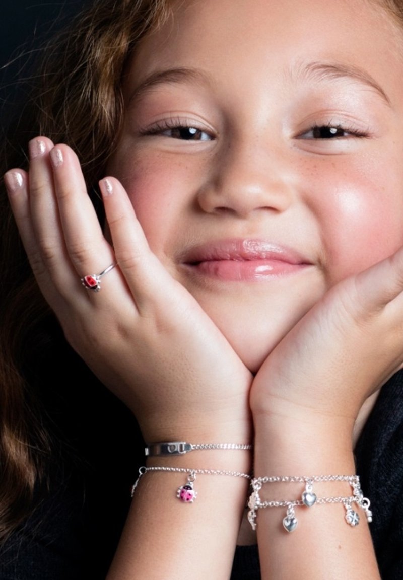 Silberne Armbänder an beiden Handgelenken zeigen Charms wie einen Marienkäfer, Herzen und Friedenssymbole. Ein schlichter silberner Ring ergänzt die Accessoires.