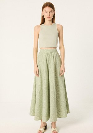 Žena, ktorá má na sebe béžový bezrukávový crop top, svetlozelenú kvetinovú maxi sukňu s vysokým pásom a béžové sandále na opätku, stojaca pred jednofarebným pozadím.
