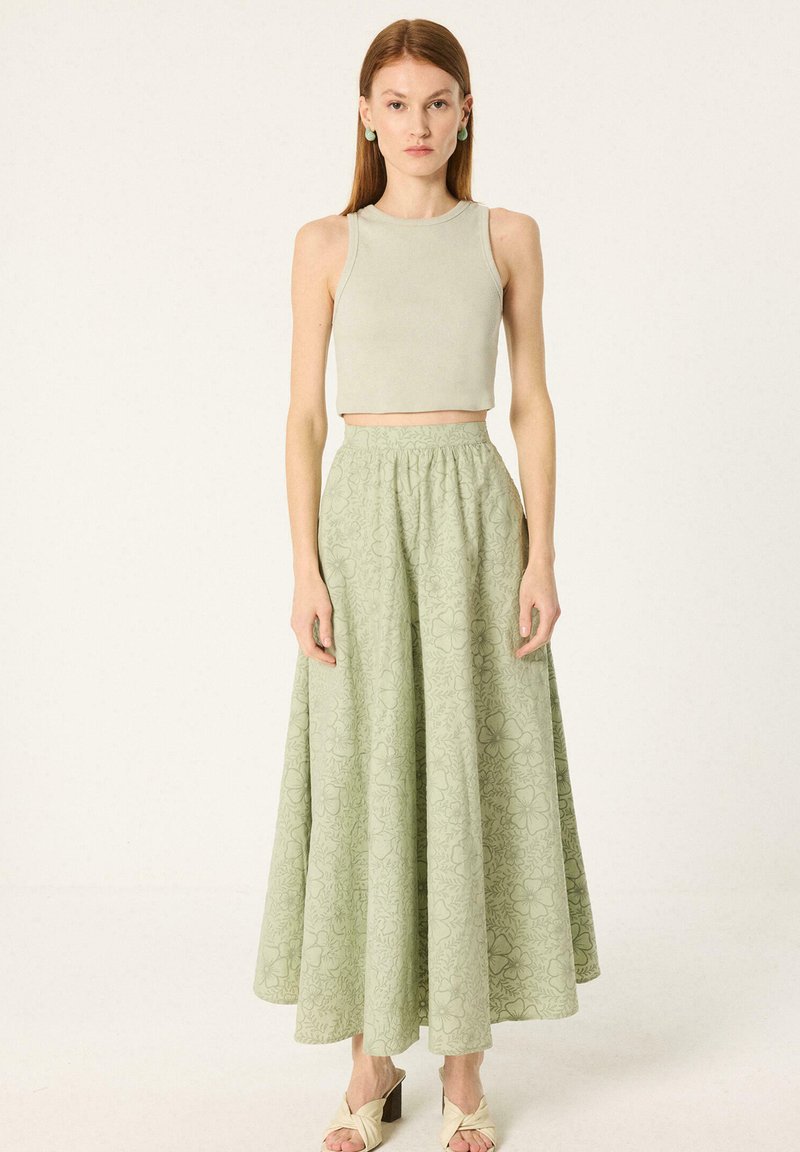Γυναίκα που φοράει μπεζ αμάνικο crop top, μακριά φούστα με λουλούδια σε ανοιχτό πράσινο χρώμα στα ψηλά, και μπεζ πέδιλα με τακούνι, στέκεται μπροστά σε απλό φόντο.