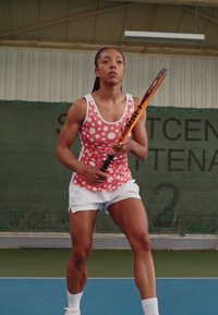 Sporty athlete in a pink polka dot tank top and white shorts, holding a tennis racket, poised on a court with a net in the background.