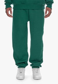 Grønne joggebukser laget av mykt stoff, med avslappet passform, ribbede mansjetter og sidelommer. Kombinert med hvite joggesko.