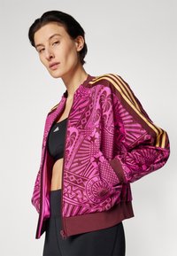 Roze bomberjack met een patroon en bruine accenten, met een ritssluiting aan de voorkant en geribde manchetten. Drie diagonale gouden strepen op de schouders.