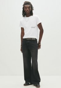 Hvid t-shirt med sort tekst, parret med brede sorte jeans og et nittet bælte. Mørke sko fuldender outfittet, med et struktureret design.