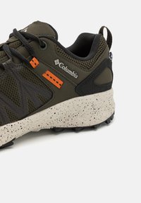 Chaussure de randonnée verte avec des accents orange et un logo Columbia. Elle présente un upper texturé, une semelle intermédiaire mouchetée et une semelle noire robuste.