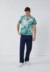 Camisa de manga corta, con cuello, en color azul verdoso con un patrón de palmeras blancas, combinada con jeans oscuros y zapatillas blancas, apoyado contra una pared blanca.