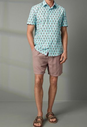 Mand iført lyseblå kortærmet skjorte med blomstermønster, lysebrune shorts og brune sandaler, stående med den ene hånd i lommen.