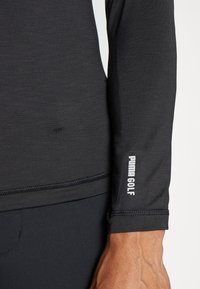 Fekete, hosszú ujjú golfpóló texturált anyagból készült, lapos varrással, és fehér "PUMA GOLF" logó található a mandzsettán. Testre szabott kialakítás sima felülettel.