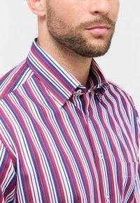 Camicia a righe nei colori rosso, bianco e blu; presenta un colletto con bottoni, una tasca sul petto e un tessuto liscio e leggero. Motivi verticali.