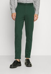 Gröna avsmalnande byxor i en texturerad tyg, med knappstängning och uppvikta fållar, i kombination med svarta snörskor.