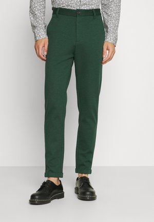 Pantalons vert en tapered en tissu texturé, avec une fermeture à bouton et des poignets retroussés, accompagnés de chaussures à lacets noires.