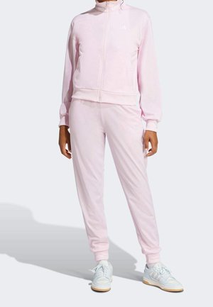 Personne portant un survêtement zippé rose pâle et des baskets blanches, debout devant un fond uni clair.