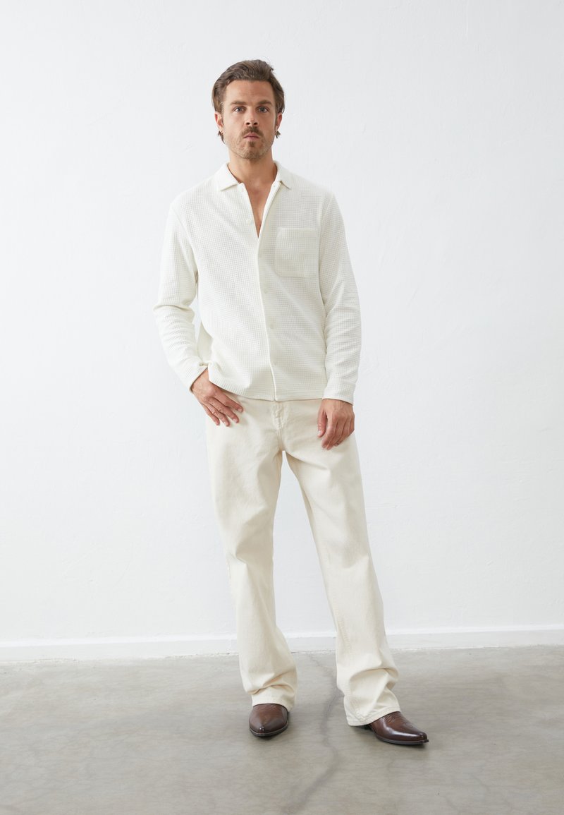Camisa de punto waffle en color blanco roto con un bolsillo en el pecho, combinada con pantalones beige claros y zapatos marrones de punta afilada, de pie sobre un suelo gris liso.