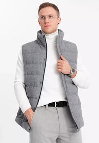 Grå pufferväst med hög krage, quiltad textur, framdragkedja och tryckknappar, bärs över en vit polo och grå byxor.