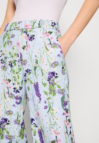 Personne portant un pantalon bleu clair à imprimé floral coloré et un haut blanc, la main reposant dans la poche du pantalon.