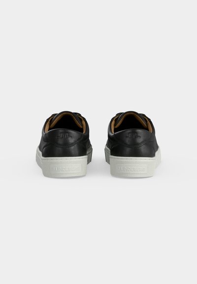 Ζευγάρι μαύρα χαμηλά sneakers με λευκές σόλες, που φαίνονται από πίσω, παρουσιάζοντας ένα λογότυπο ανάγλυφο στην περιοχή της φτέρνας.