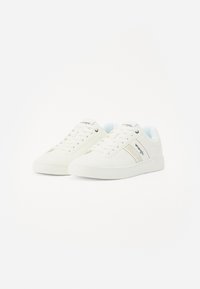Baskets en toile blanches avec une semelle en caoutchouc, présentant une texture lisse, un bout arrondi et une bande latérale en accent. Détails des lacets et du logo visibles.