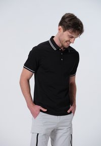 Camisa polo negra con ribete blanco en el cuello y las mangas, presenta una tapeta con tres botones y está hecha de un suave material de algodón.
