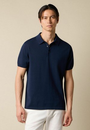 Un jeune homme se tient face à l'avant, portant un polo bleu marine à manches courtes et un pantalon blanc, sur un fond beige uni.