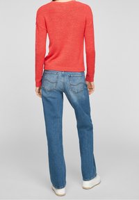 Pull en tricot rouge texturé avec un col rond, associé à un jean en denim bleu clair à coupe droite, doté de poches arrières et de coutures légères.