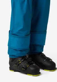 Zwarte skibotten met drie verstelbare gespen en groene accenten, gecombineerd met blauwe waterdichte broek die bij de enkels is gathered.