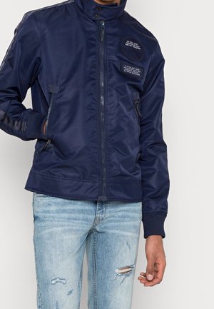 Personne portant une veste bomber bleu marine zippée avec deux écussons de texte, et un jean bleu clair déchiré, se tenant devant un fond blanc uni.