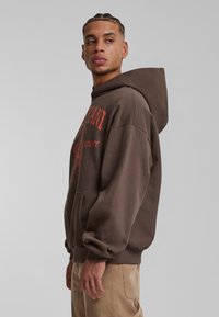 Sudadera con capucha marrón y bolsillo frontal, con texto gráfico y diseño en rojo. Corte holgado con puños y dobladillo acanalados, combinada con pantalones beige.