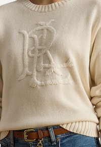Pull en tricot crème avec un logo brodé texturé au centre, encolure ronde et poignets et ourlet côtelés.