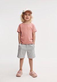 Rosa bomulls t-skjorte med en liten rød logo, stripete shorts i lys grå og hvit, og rosa klogs med dekorative detaljer.