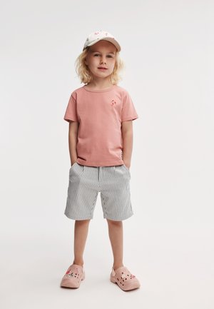 T-shirt en coton rose avec un petit logo rouge, short rayé en gris clair et blanc, et sabots roses avec des accents décoratifs.