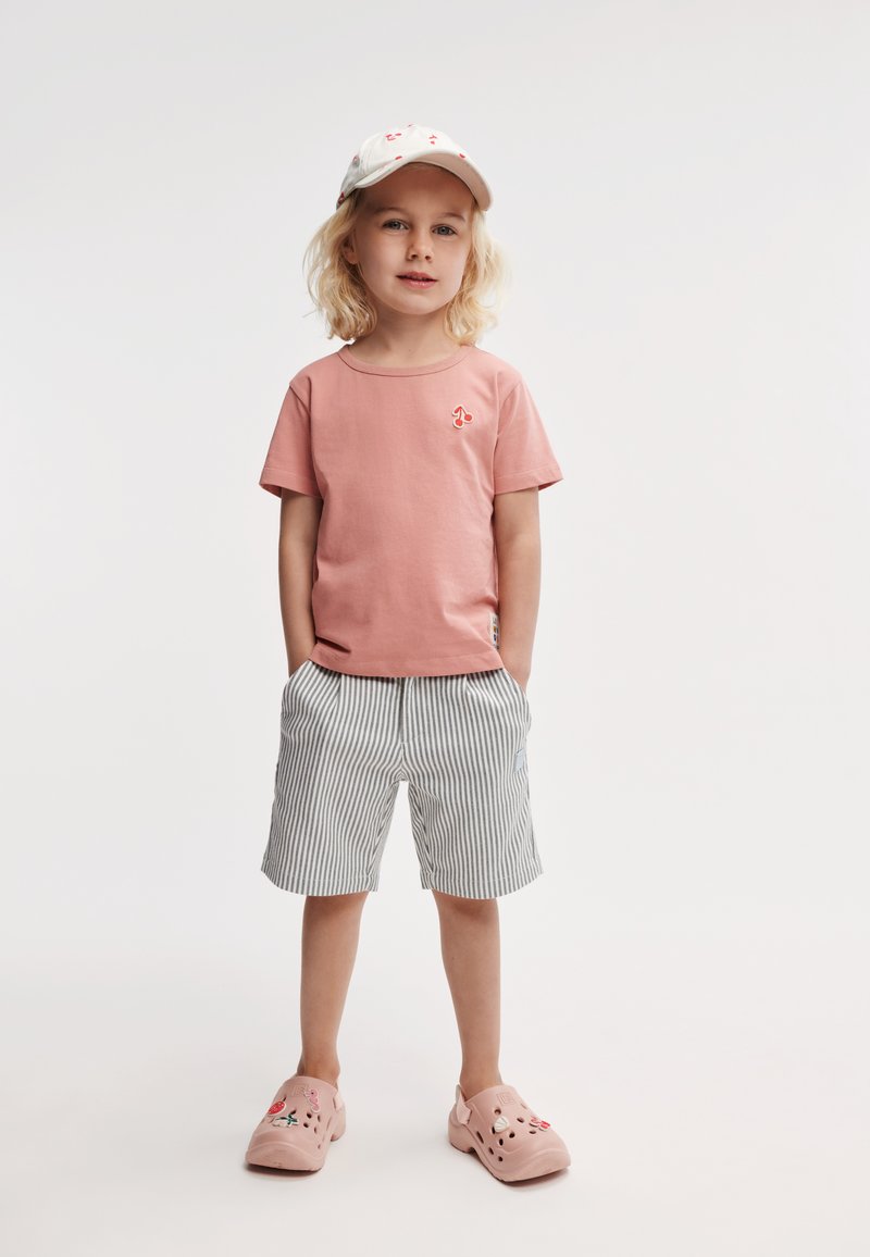 Rosa bomulls t-skjorte med en liten rød logo, stripete shorts i lys grå og hvit, og rosa klogs med dekorative detaljer.