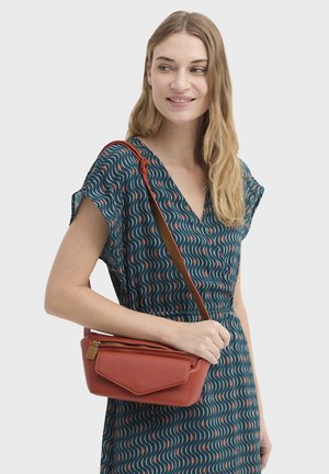 Kvinde smilende, iført en blå mønstret kjole, der holder en lille tekstureret skråtaske i rustfarve med en forlommelomme med lynlås.