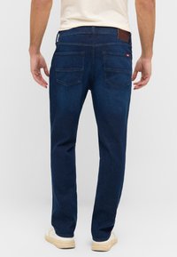 Jeans de mezclilla de color azul oscuro con corte recto, que cuentan con dos bolsillos traseros y un parche de cuero marrón en la cintura. Combinados con zapatillas blancas.