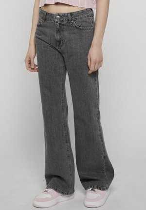 Oseba, ki nosi široke črne jeans hlače z visokim pasom in bledim učinkom, kombinirane z roza in belimi supergami, stoji pred enostavnim svetlim ozadjem.