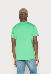T-shirt verde a maniche corte realizzato in morbido cotone, con un classico scollo rotondo e una texture liscia e uniforme. Indossato con jeans blu.