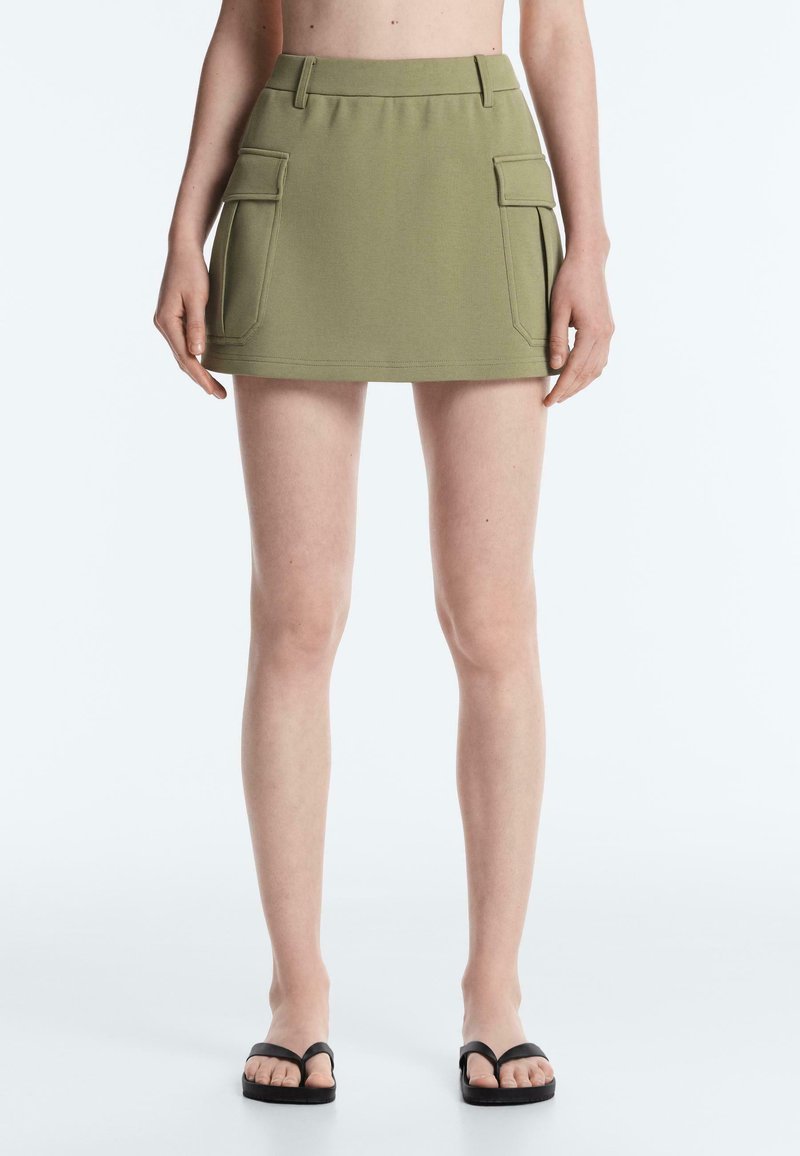 Femme portant une mini-jupe vert olive avec des poches à rabat devant et des tongs noires, debout devant un fond blanc uni.