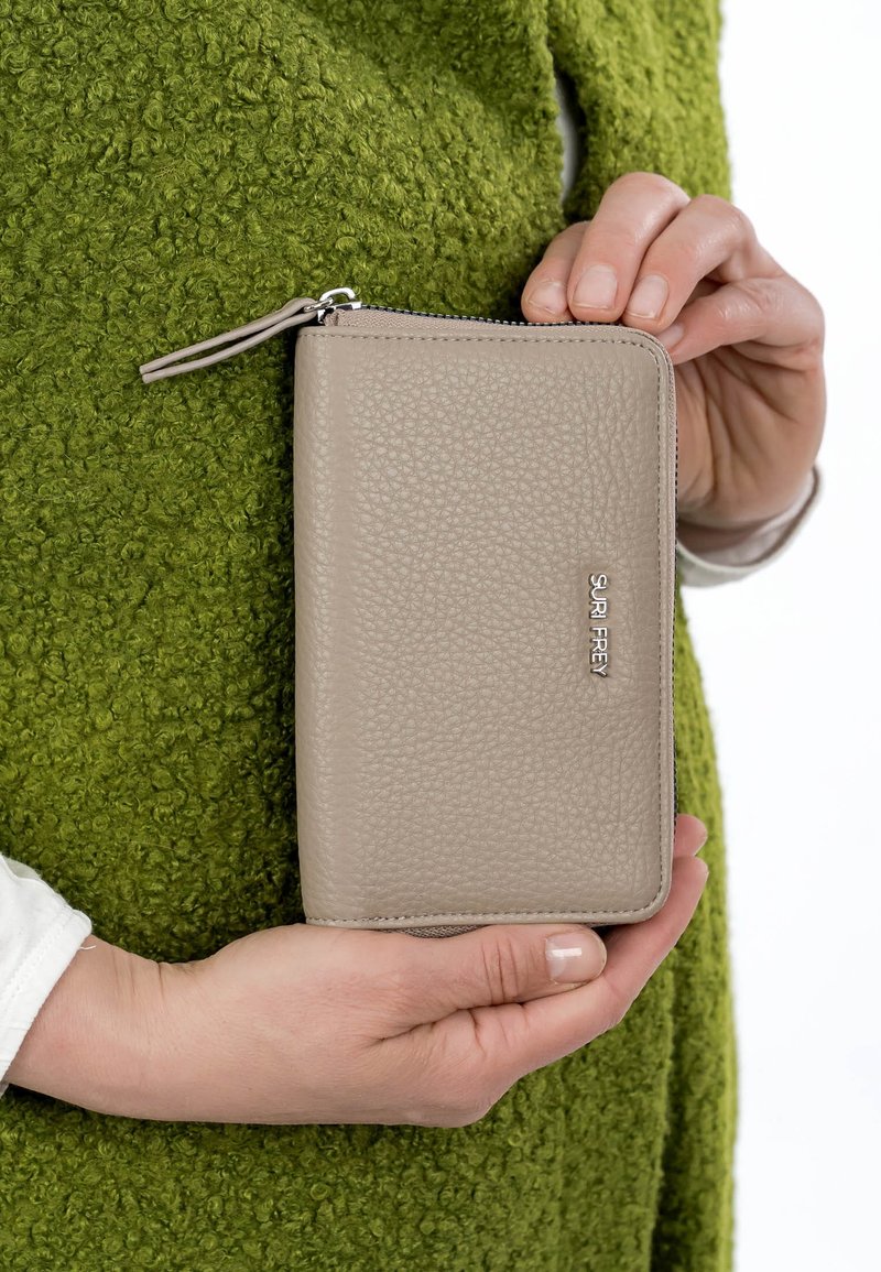 Hände halten eine beige strukturierte Leder-Brieftasche mit Reißverschluss und dem Logo "SURI FREY" vor einem grünen, flauschigen Stoffhintergrund.