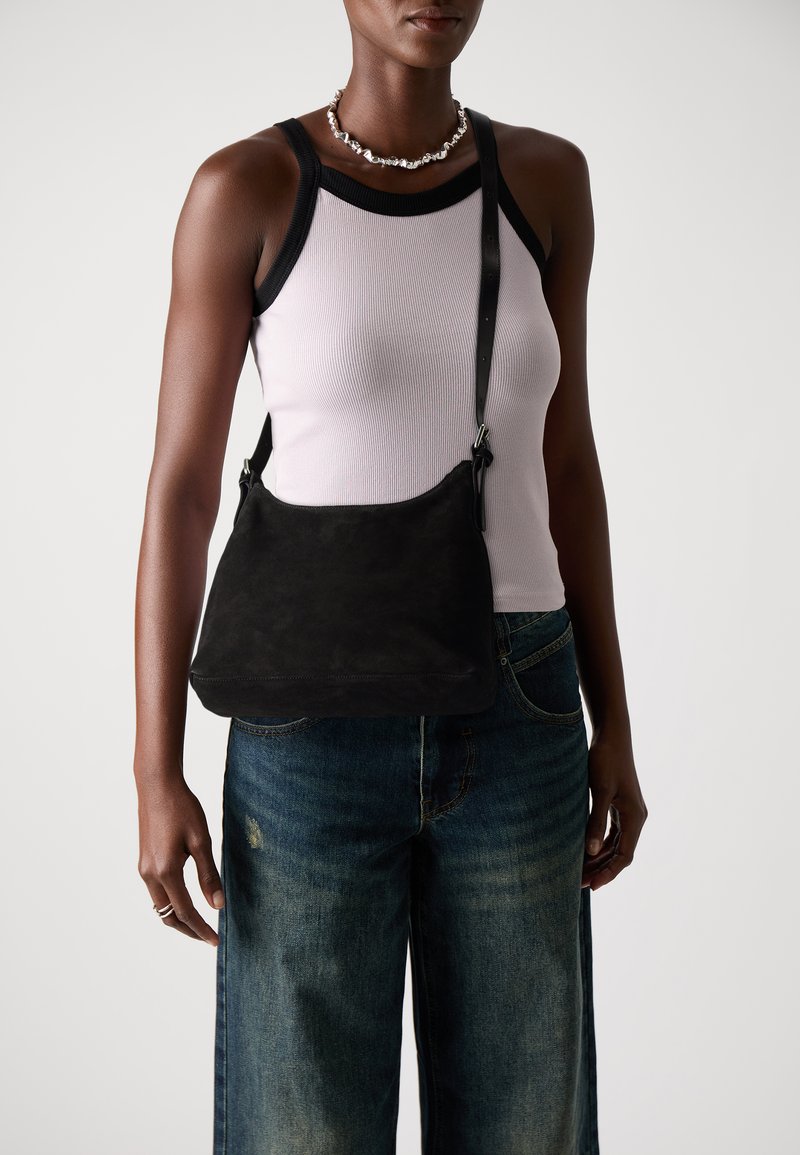 Zwarte suède schoudertas met een zachte, gestructureerde vorm; verstelbare schouderriem; minimale hardware; gecombineerd met een licht tanktop en wijde jeans.