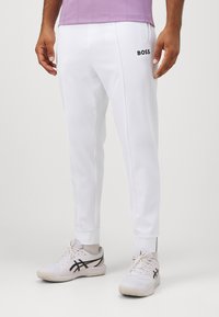 Witte sportbroeken met geribbelde boorden, voorzien van een zwart "BOSS" logo op de linker bovenbeen. Lichte stof, taps toelopend ontwerp, gecombineerd met witte sneakers.