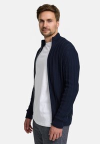 Marineblaue, gerippte Strickstrickjacke mit Stehkragen, durchgehendem Reißverschluss und langen Ärmeln. Kombiniert mit einem weißen T-Shirt und grauen Hose.
