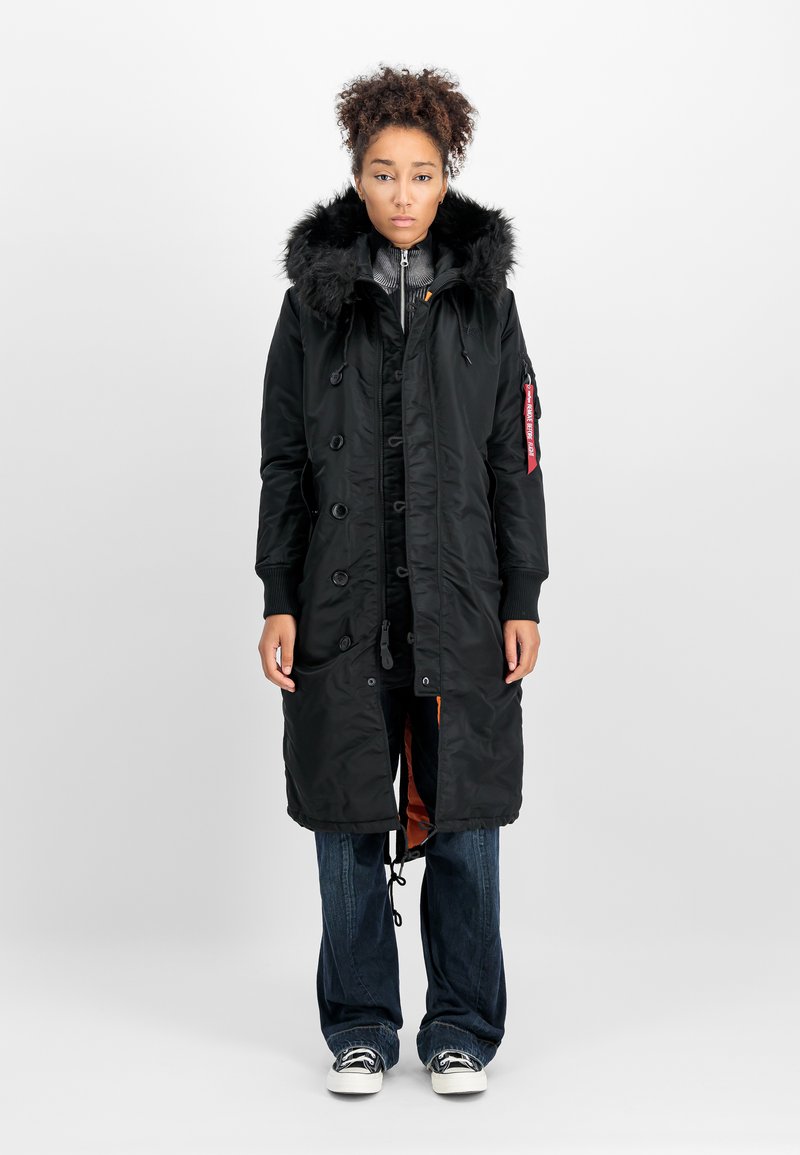 Vrouw die staat in een lange zwarte winterjas met een met bont gevoerde capuchon, gelaagd over donkere spijkerbroeken en zwart-witte sneakers, tegen een effen achtergrond.