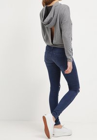 Sudadera gris con capucha y espalda descubierta, combinada con jeans ajustados azul oscuro con rodillas desgastadas y zapatillas blancas con suelas texturizadas.