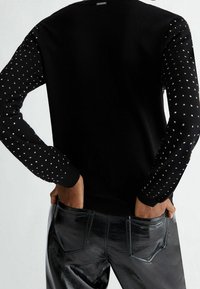 Suéter de punto negro con mangas largas que presenta un patrón texturizado de pequeños cuadrados. Cuello de canalé y dobladillo ajustado. Se lleva con pantalones negros brillantes.