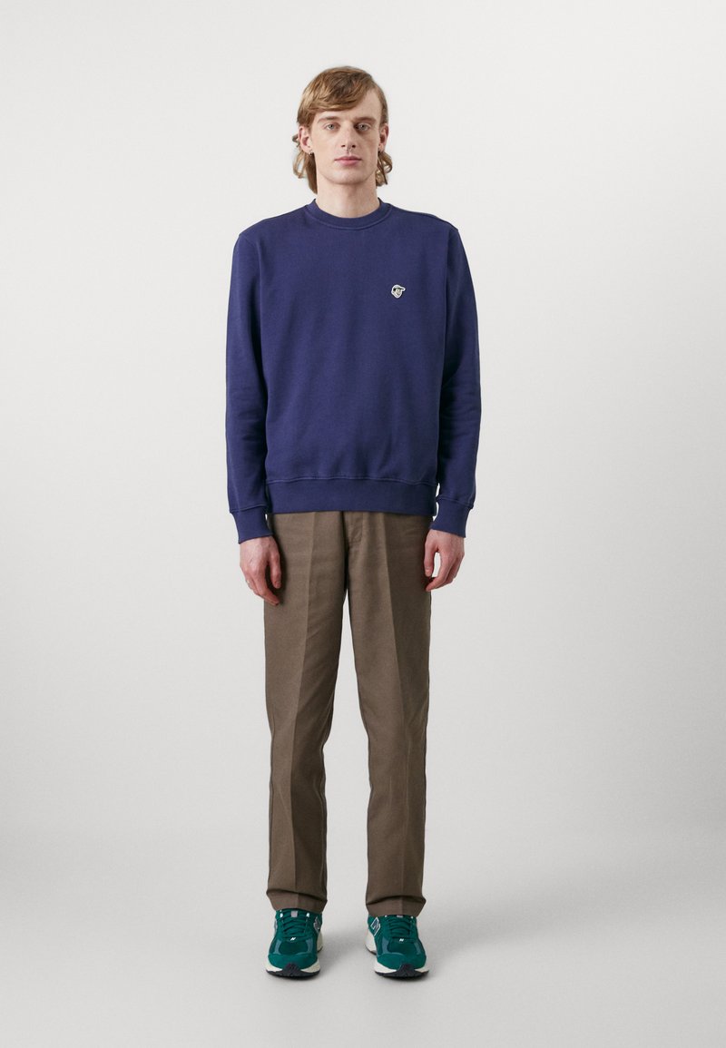 Felpa viola con collo rotondo e piccolo logo, abbinata a pantaloni marroni a gamba affusolata e sneakers verdi. Tessuto morbido e vestibilità rilassata.