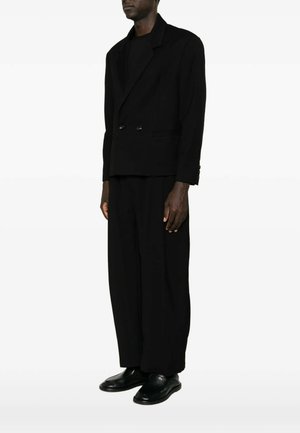 Man draagt een loszittende zwarte double-breasted blazer, wijde zwarte broek, zwart overhemd en zwarte leren schoenen en staat zijwaarts.