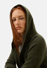 Sweat à capuche vert foncé avec texture côtelée, présentant une fermeture éclair à l'avant et une coupe décontractée. L'arrière-plan est blanc uni pour le contraste.