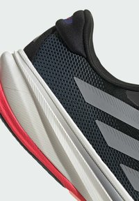 Zapatilla deportiva con parte superior de malla negra, rayas diagonales grises y una suela texturizada blanca y roja. Presenta un collar acolchado para mayor comodidad.