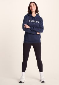 Navy hooded sweatshirt with “TOG24” lettering, cotton blend material, adjustable drawstrings, worn with black leggings and white sneakers.
