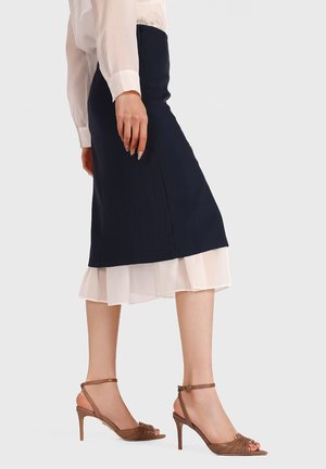 Donna che indossa una gonna nera sopra un vestito bianco con sandali marroni con tacco alto, in piedi su uno sfondo semplice.